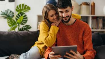 Pareja heterosexual sentada en el sofá de su casa viendo algo en su tablet.