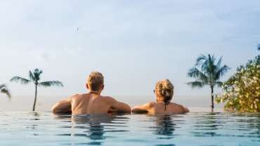 Pareja heterosexual dentro de una alberca de borde infinito frente a una hermosa vista con palmeras grandes.