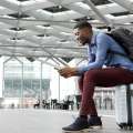 Hombre negro sentado sobre su propia maleta en un aeropuerto usando su celular
