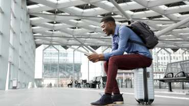 Hombre negro sentado sobre su propia maleta en un aeropuerto usando su celular