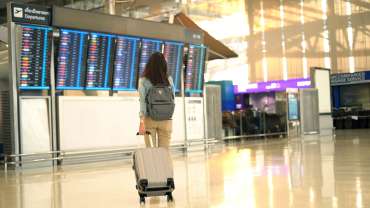 Mujer sosteniendo una maleta frente al panel del aeropuerto.